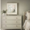 Botanical canvas print featuring delicate wildflowers in sage green and blush pink tones, displayed in white float frame above cream dresser in neutral bedroom setting with white bedding and floral area rug