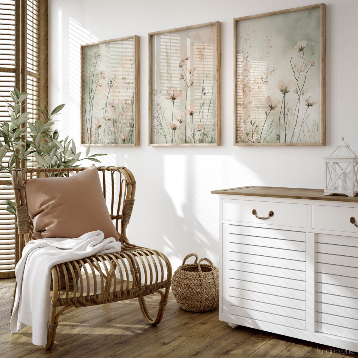 Set of three framed botanical prints featuring delicate wildflowers and grasses in neutral tones, displayed above a white coastal sideboard next to a rattan chair with natural light streaming through wooden shutters in a bright boho-style living room