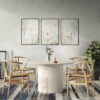 Set of three framed botanical prints featuring delicate wildflowers and dried grasses in neutral beige tones, displayed above a modern concrete dining table with wooden wishbone chairs in a contemporary dining room with gray walls