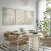 Set of three framed botanical prints featuring delicate wildflowers and grasses in soft neutral tones, displayed above a round natural wood dining table with woven rattan chairs and green ceramic dishes in a bright modern dining room with large windows and plants
