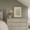 Framed botanical print featuring delicate wildflowers and cosmos in soft pink and beige watercolor tones, hanging above an ornate cream French provincial nightstand with cherry blossoms in a gold vase, styled in an elegant bedroom with sage green walls