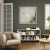 Large botanical canvas print featuring sage green wildflowers in white float frame, displayed above white bookshelf in modern living room with beige armchair, hardwood floors, and sage green accent wall