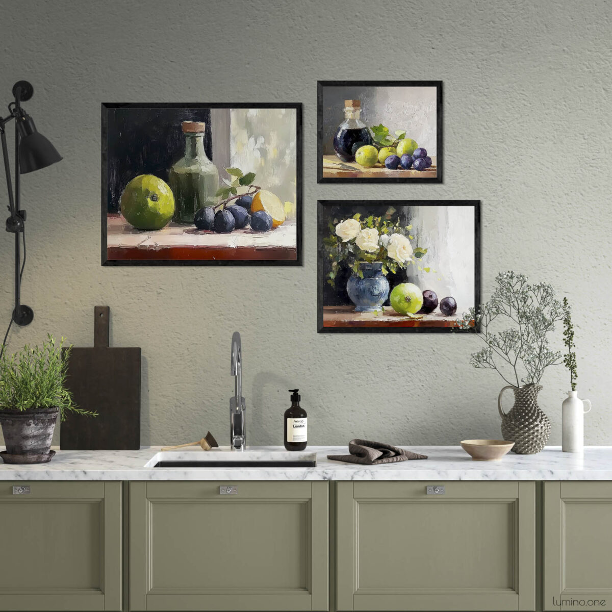 Three framed still life oil painting prints displayed as gallery wall above kitchen counter featuring fruits, flowers in blue vase, and glass bottles in classic painterly style, shown in modern kitchen with white marble countertop and sage green cabinets